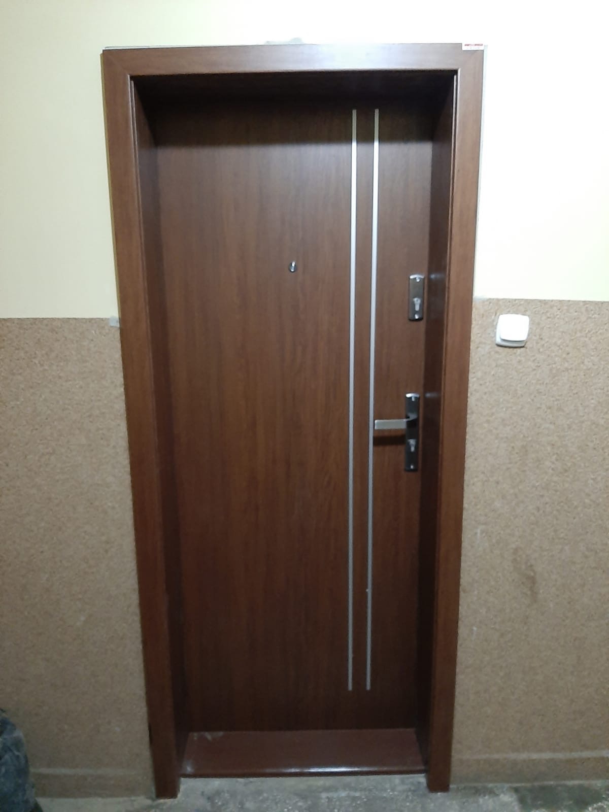 Brązowe drzwi wejściowe z dwoma pionowymi, srebrnymi paskami, wizjerem i zamkiem, zamontowane w korytarzu z beżowymi ścianami o ziarnistej fakturze.