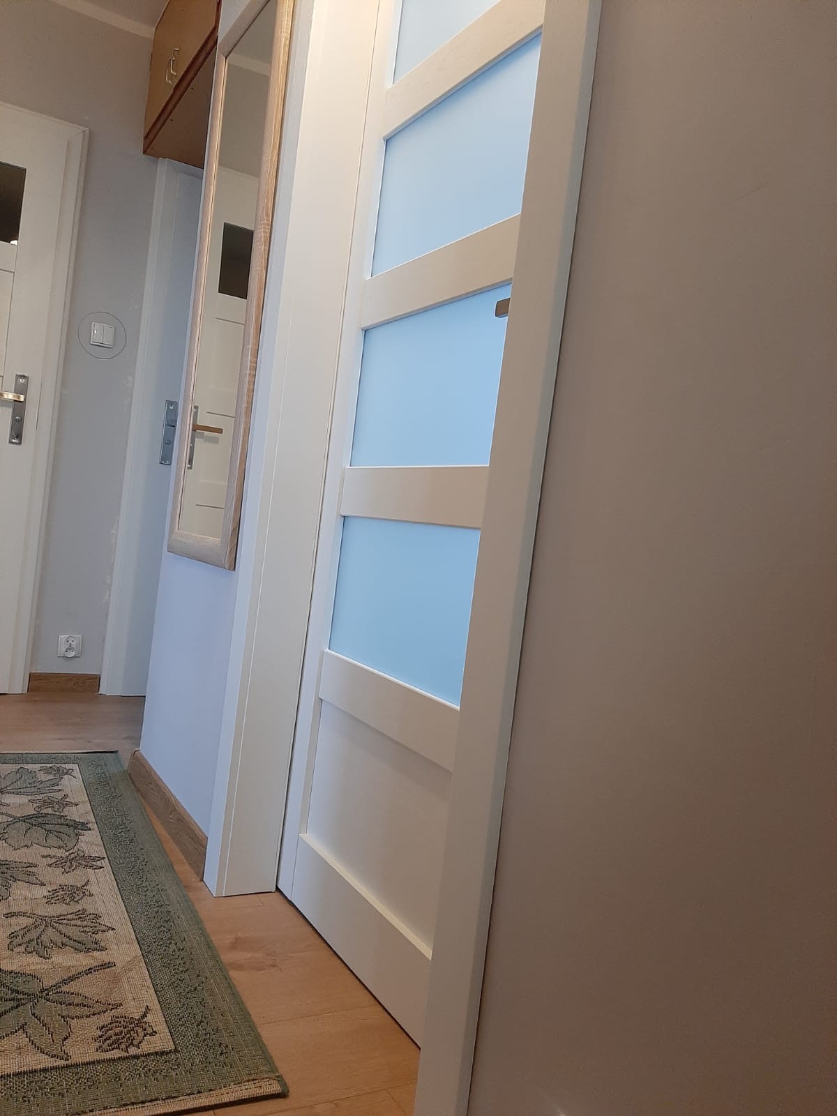 Białe, częściowo przeszklone drzwi wewnętrzne z trzema poziomymi panelami z matowego, błękitnego szkła, widoczne w korytarzu z dywanem w odcieniach zieleni i beżu.