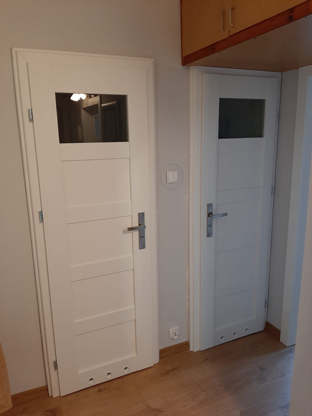 Dwa białe skrzydła drzwiowe z szybami w górnej części, widoczne w korytarzu z drewnianą podłogą i szafkami pod sufitem, jedno skrzydło z trzema otworami wentylacyjnymi u dołu.