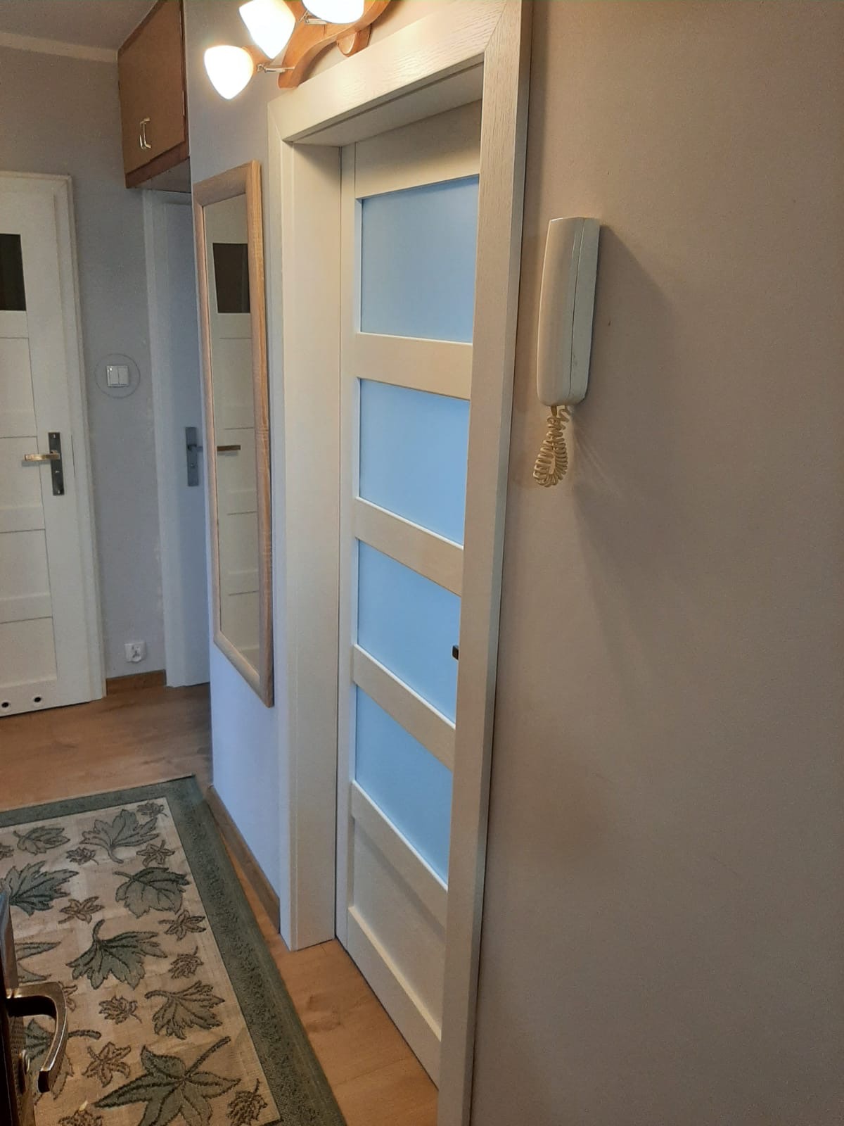 Białe drzwi wewnętrzne z czterema panelami wypełnionymi błękitnym szkłem, widoczne z boku w korytarzu, obok domofon i lustro w drewnianej ramie.