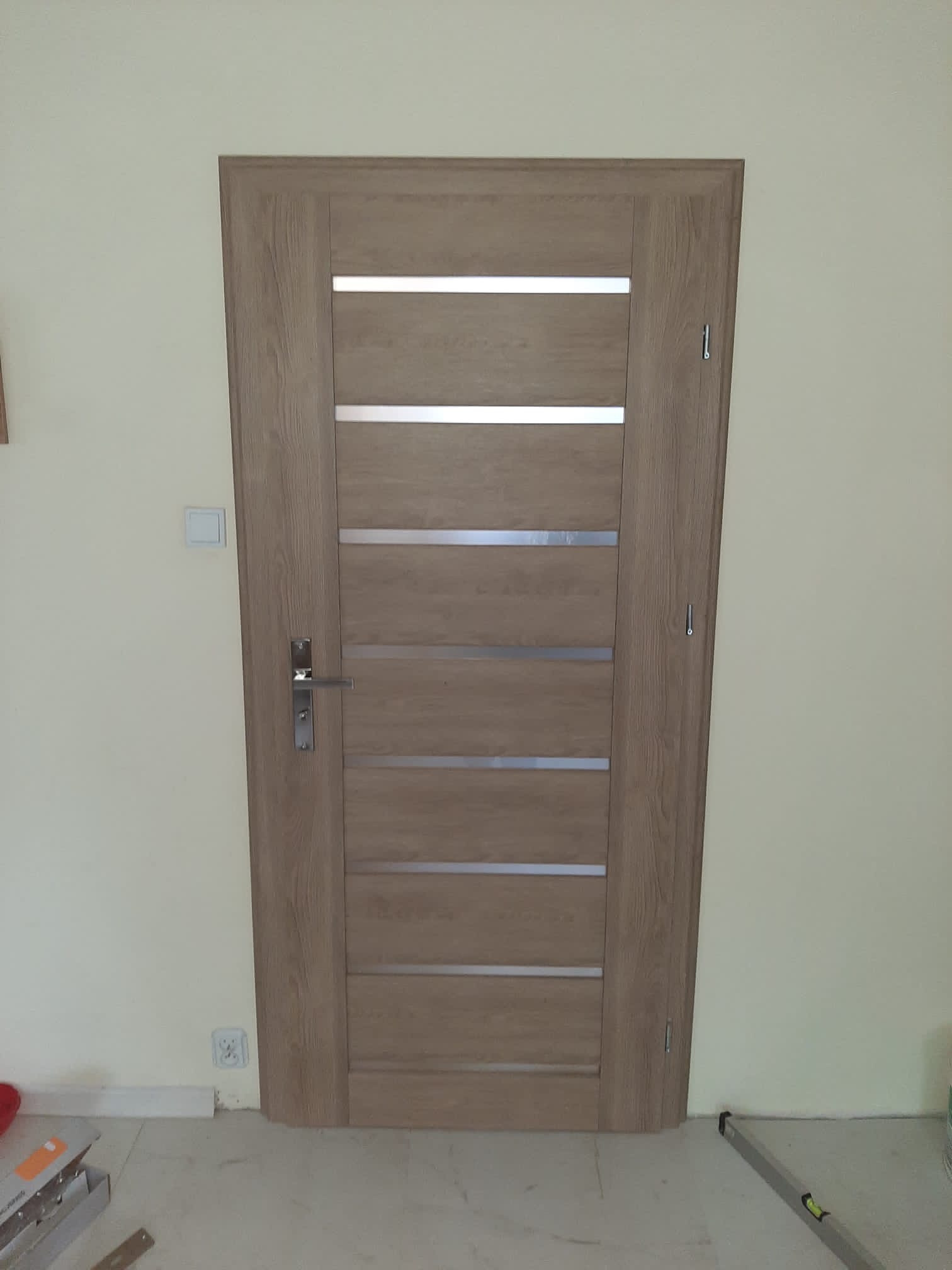 Brązowe drzwi wewnętrzne z poziomymi, srebrnymi wstawkami, zamontowane w ścianie, widoczne elementy wykończeniowe podłogi i poziomica.