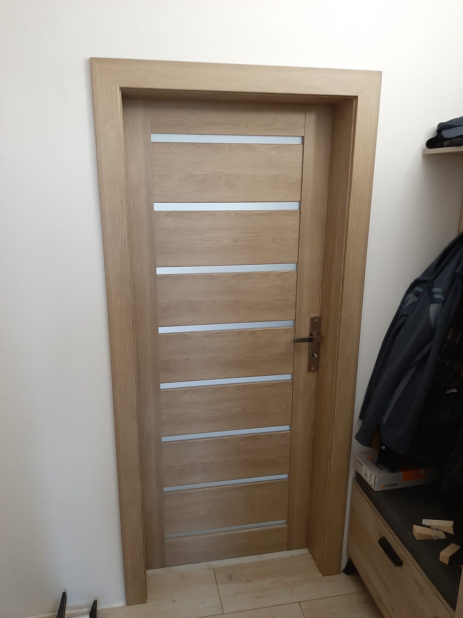 Jasne, drewniane drzwi wewnętrzne z poziomymi, matowymi przeszkleniami, zamontowane w ościeżnicy. Na podłodze widoczne jasne płytki. Część garderoby i ubrania widoczne z prawej strony.