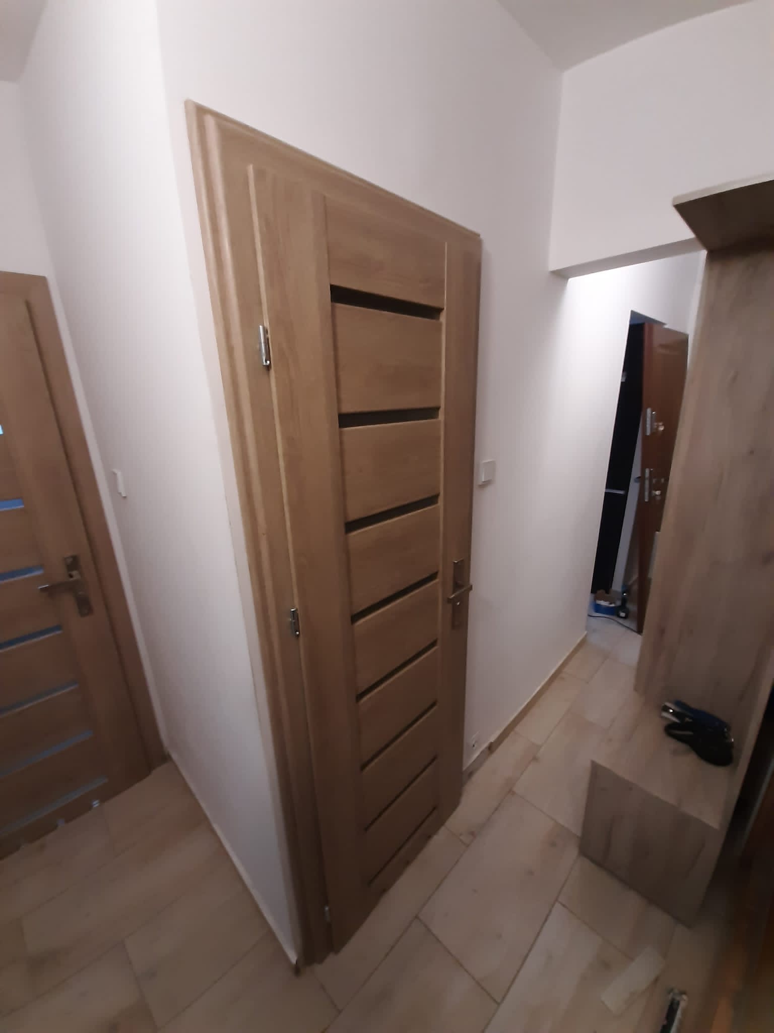 Nowoczesne drzwi wewnętrzne z poziomymi przeszkleniami w jasnym drewnie, zamontowane w korytarzu z drewnianą podłogą i szafką na buty.