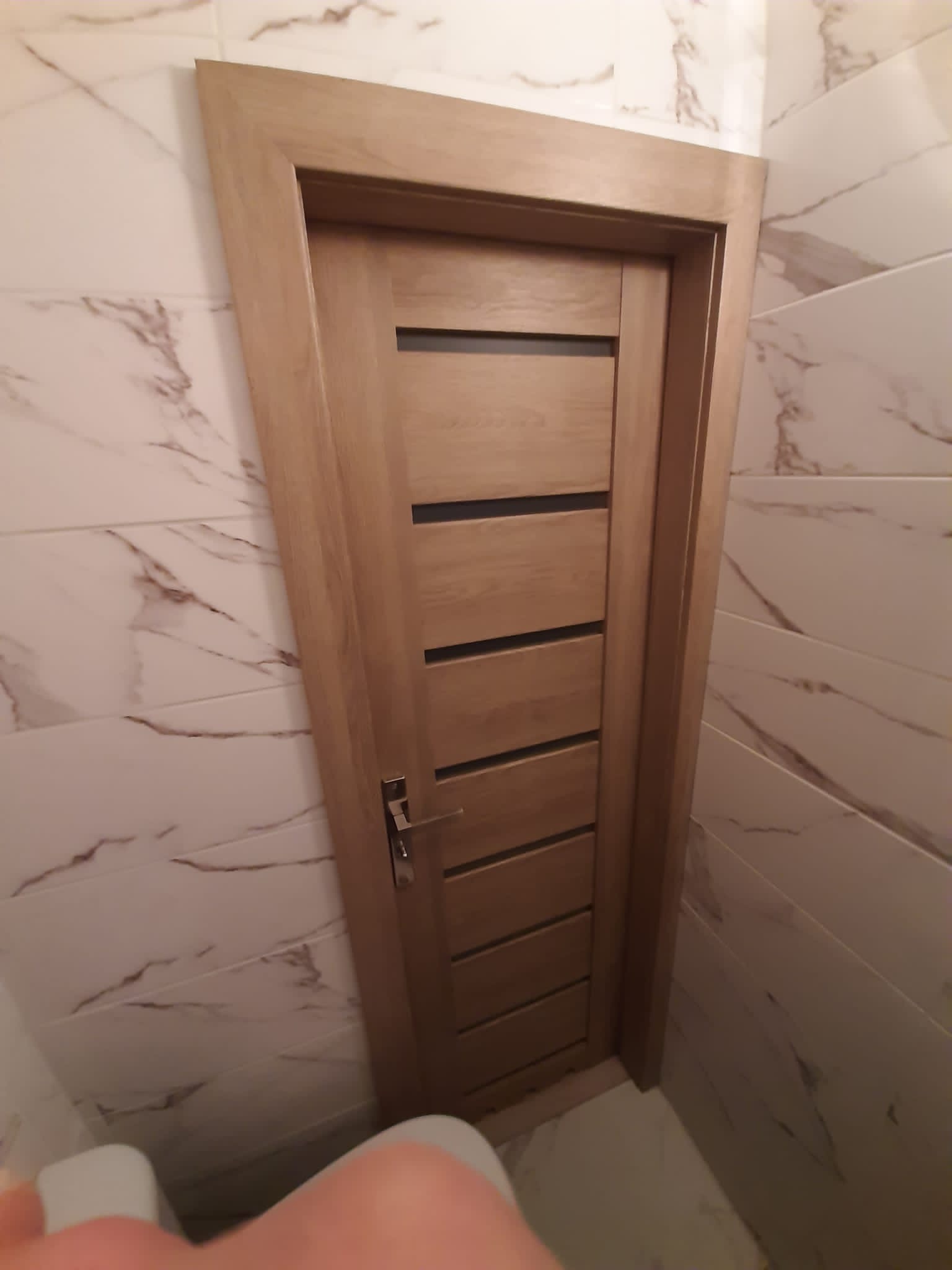 Zamknięte drzwi wewnętrzne w drewnianej ościeżnicy z poziomymi, ciemnymi wstawkami, zamontowane w łazience wyłożonej białymi płytkami z marmurowym wzorem.