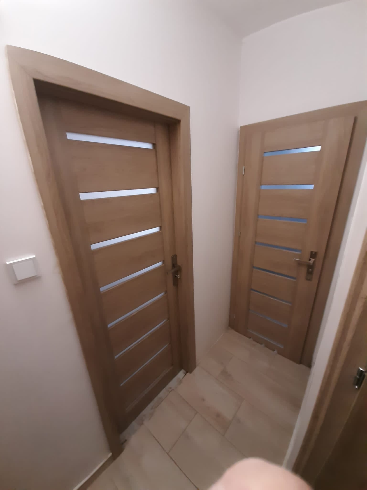 Dwa drewniane skrzydła drzwiowe z poziomymi, prostokątnymi przeszkleniami, zamontowane w jasnym korytarzu z płytkami imitującymi drewno.