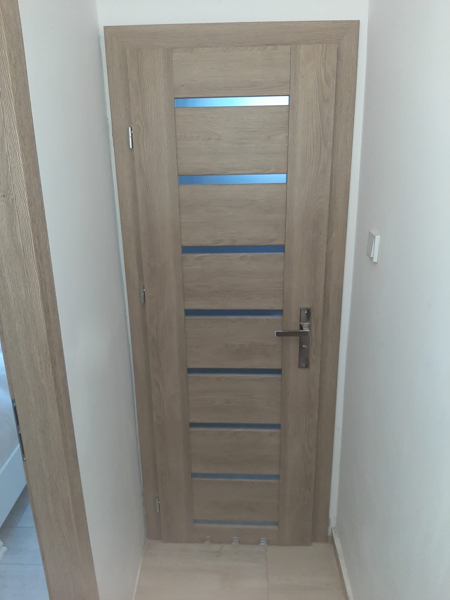 Zamknięte, drewniane drzwi wewnętrzne z poziomymi, niebieskimi wstawkami, widoczne z bliska w korytarzu z białymi ścianami.