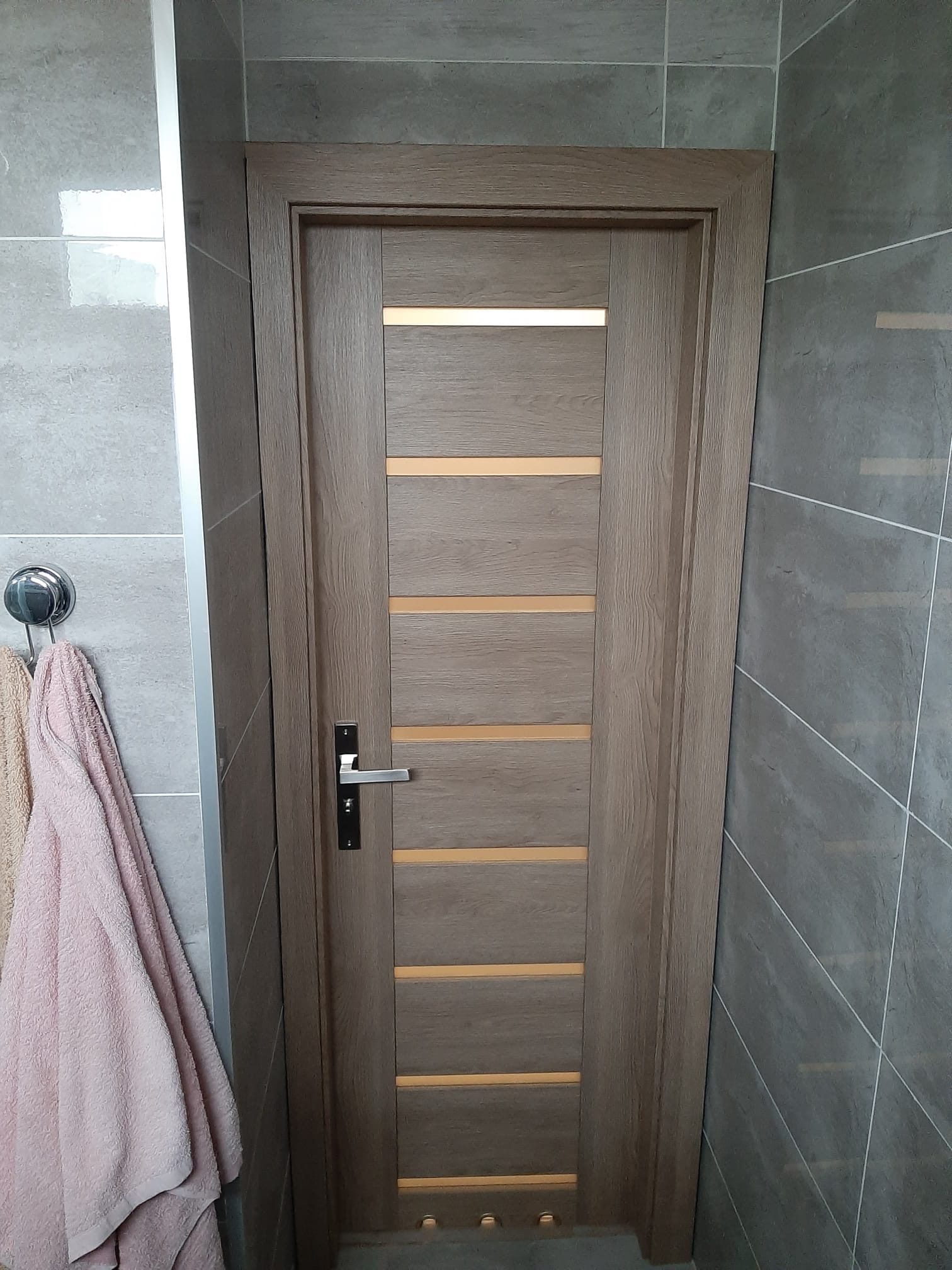 Zamknięte, drewniane drzwi wewnętrzne z poziomymi, jasnymi wstawkami w łazience z szarymi płytkami, z widocznym wieszakiem na ręczniki po lewej stronie.