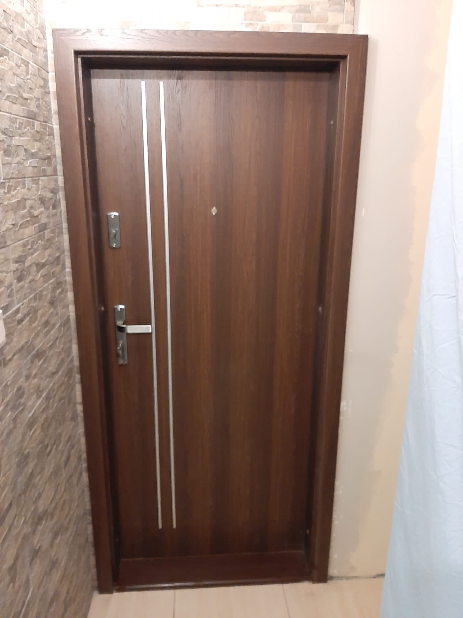 Ciemnobrązowe drzwi wejściowe z dwoma pionowymi, srebrnymi paskami, zamontowane w korytarzu wykończonym kamienną okładziną na jednej ścianie i gładką, beżową ścianą po drugiej stronie.