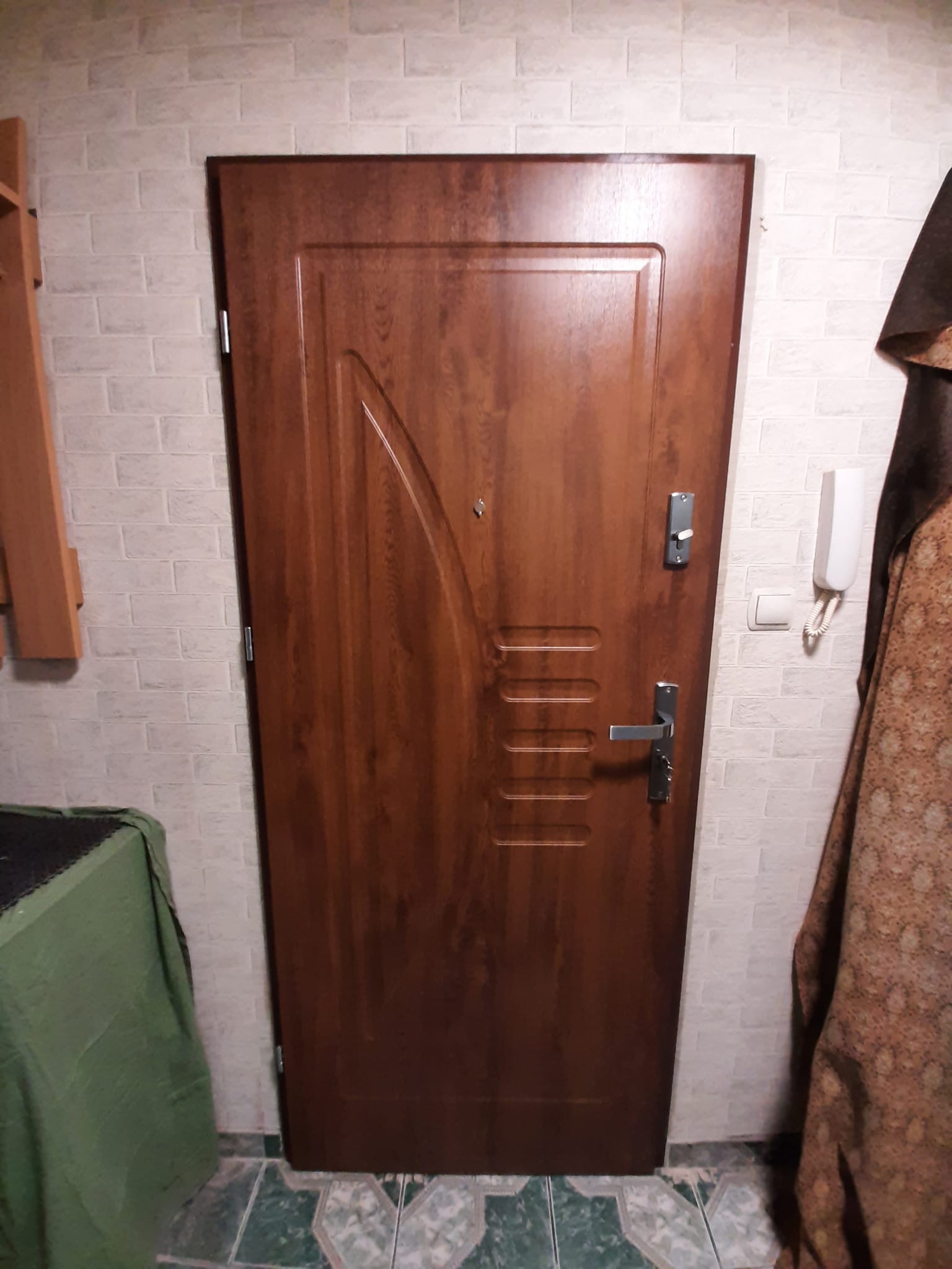 Brązowe drzwi zewnętrzne z panelem dekoracyjnym i wizjerem zamontowane w korytarzu z białą cegłą na ścianie, widoczny domofon i wieszak.