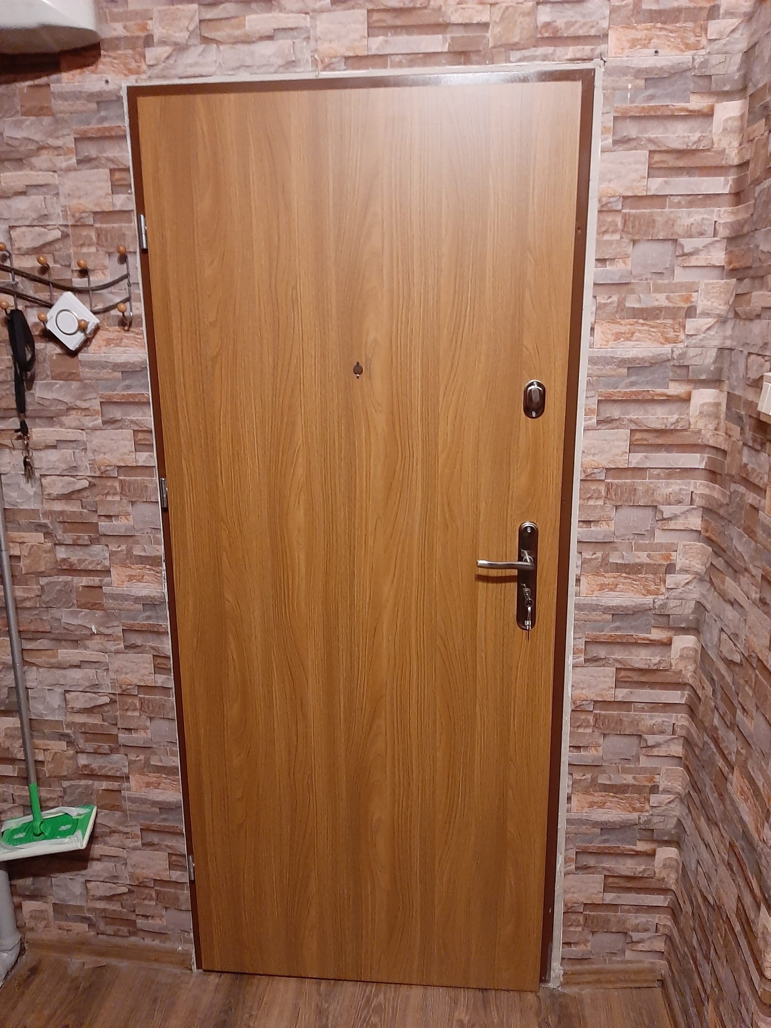 Jasnobrązowe drzwi wejściowe z wizjerem i zamkiem, na tle ściany wyłożonej kamienną okładziną w odcieniach brązu i beżu. W rogu widoczny mop.