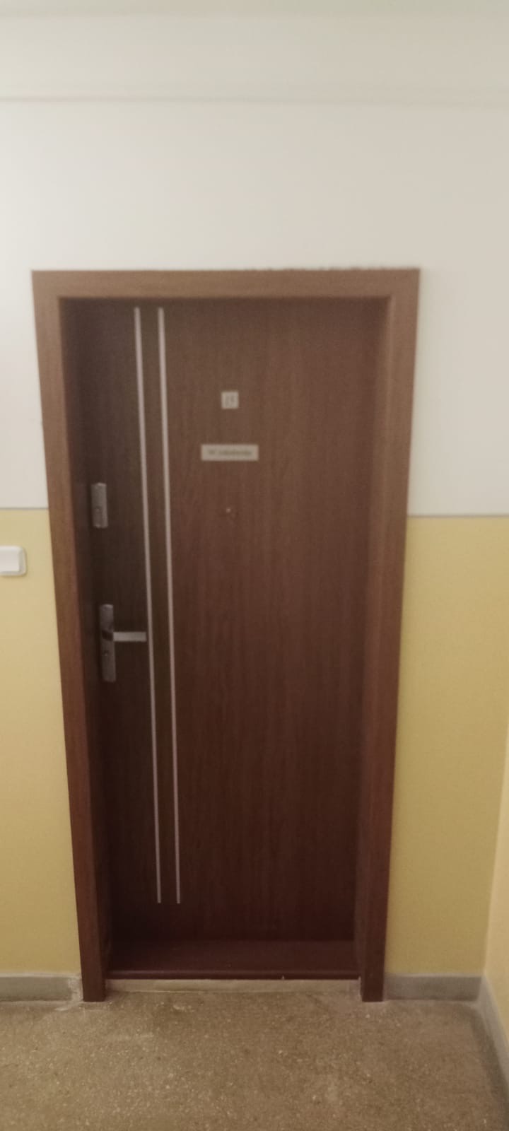 Brązowe drzwi wewnętrzne z dwoma pionowymi, białymi paskami, zamontowane w ścianie pomalowanej na biało i żółto, widoczne zamek i klamka.