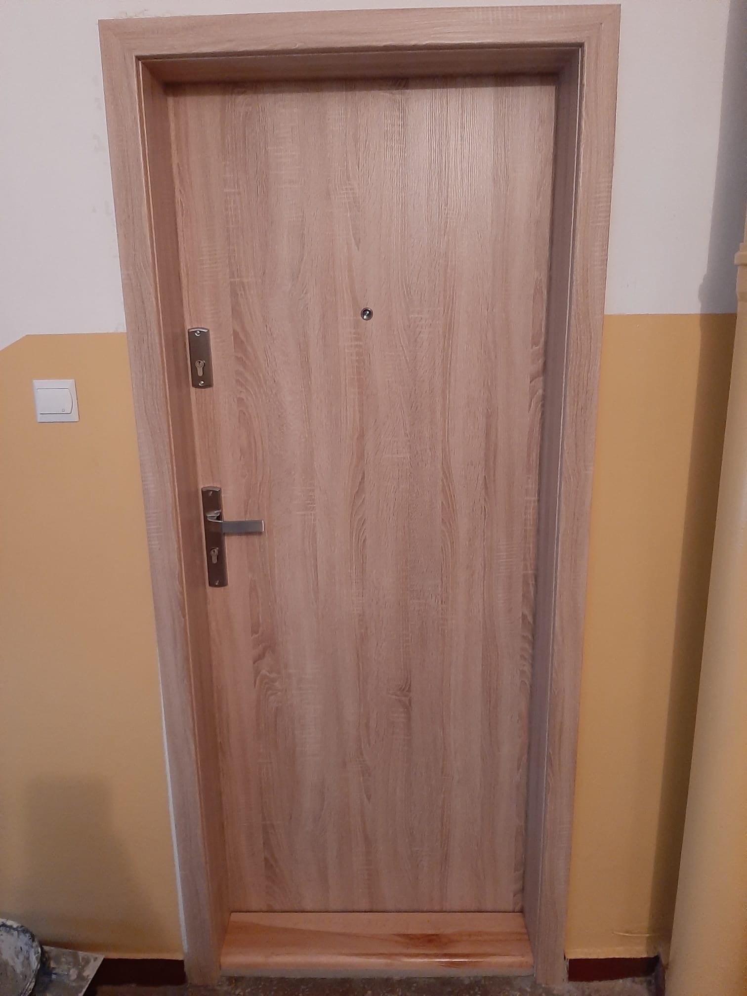 Jasne, drewnopodobne drzwi z wizjerem i srebrną klamką, zamontowane w żółtej ścianie z widocznym włącznikiem światła i fragmentem rury.