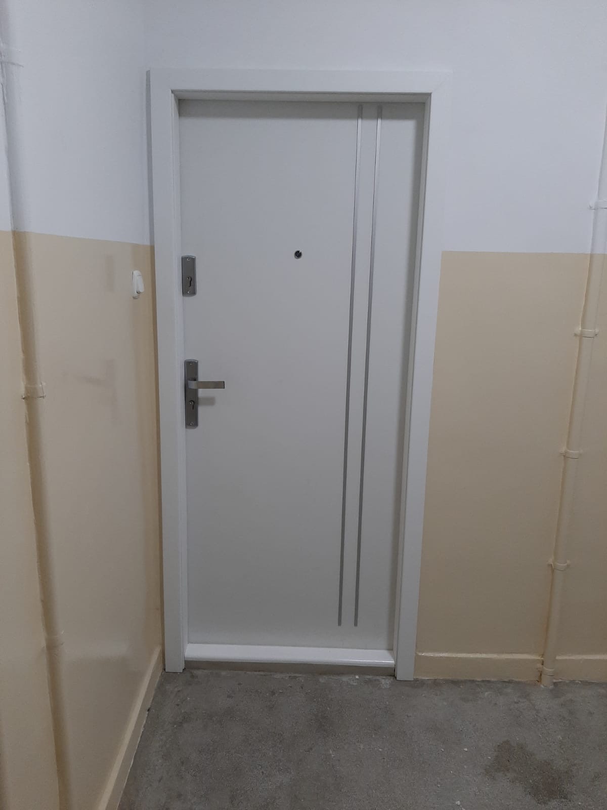 Białe drzwi wejściowe z dwoma pionowymi wgłębieniami, zamontowane w bloku w Grudziądzu, widoczne zamek i wizjer.