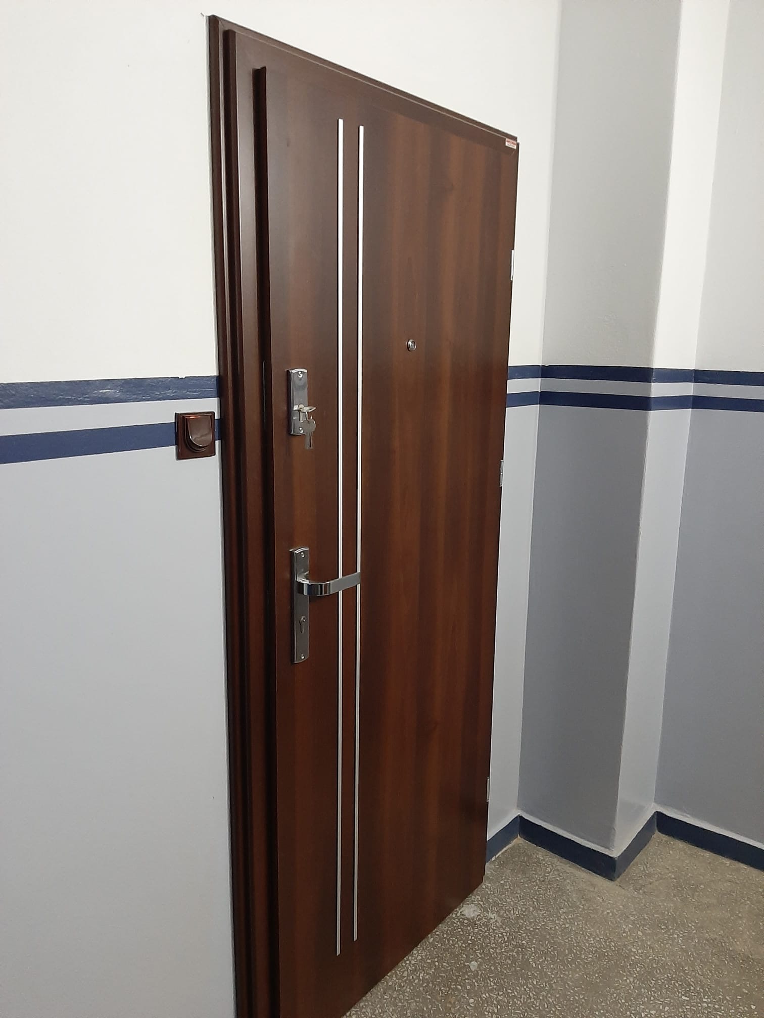 Ciemnobrązowe drzwi zewnętrzne z trzema pionowymi, srebrnymi paskami i wizjerem zamontowane w szarej klatce schodowej z niebieskim paskiem na ścianie.