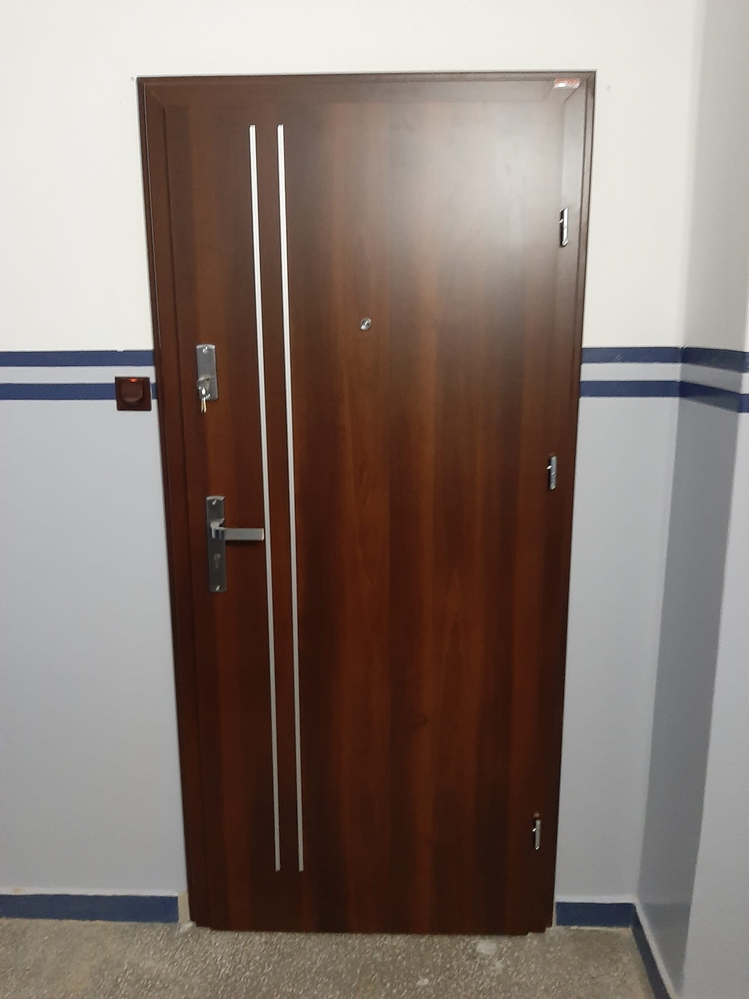 Brązowe drzwi wejściowe z dwoma srebrnymi paskami, zamkiem z kluczem i wizjerem, zamontowane w białej ścianie z niebieskim paskiem dekoracyjnym.