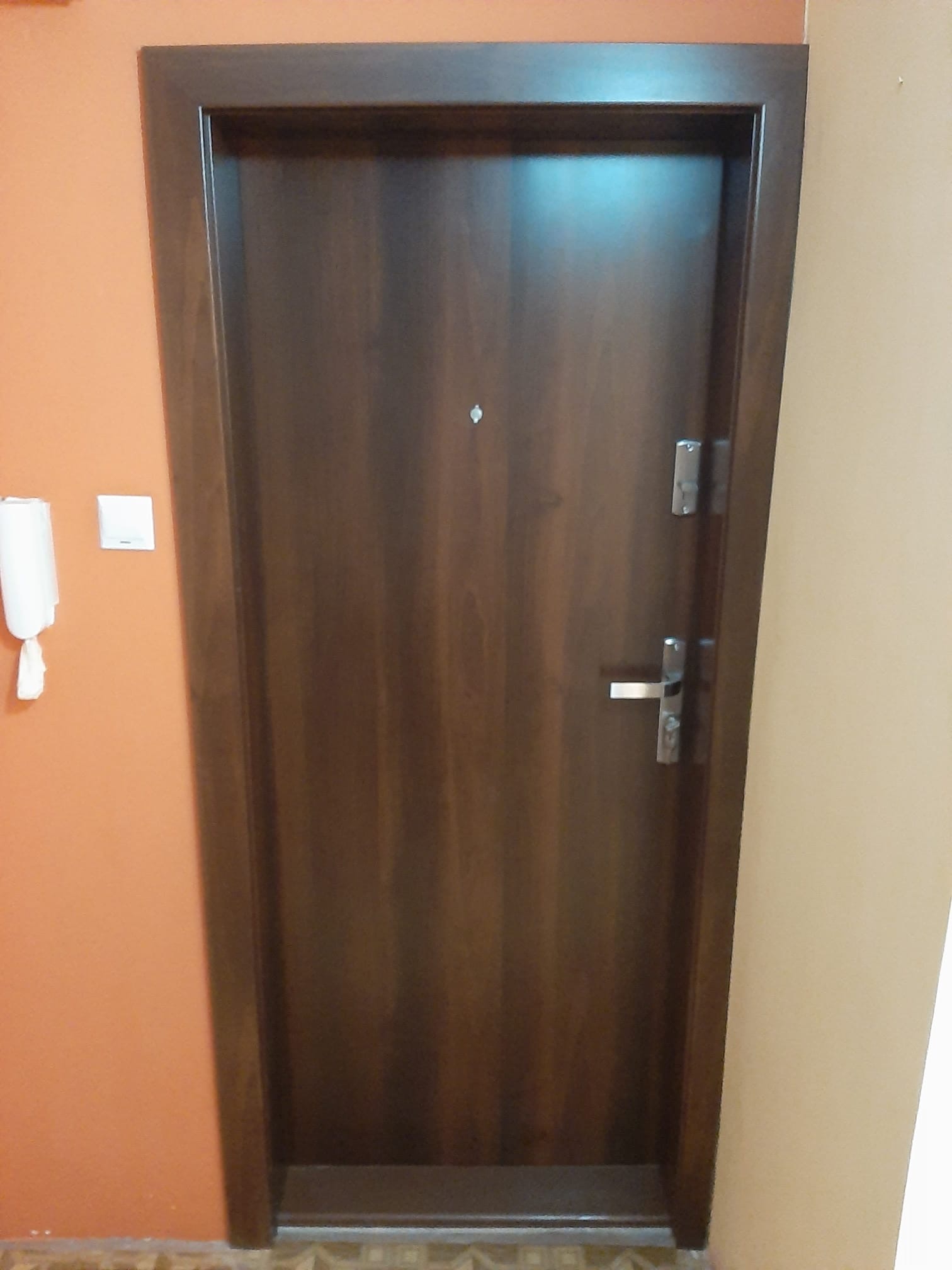 Ciemnobrązowe drzwi z wizjerem i srebrną klamką, widoczne na tle pomarańczowej i beżowej ściany, z zamontowanym domofonem po lewej stronie.