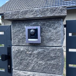 Electronic Team Mariusz Starzycki - Zamontowany wideodomofon IP Hikvision na szarym, kamiennym słupku ogrodzenia, widoczny fragment furtki i domu z szarą dachówką w tle.