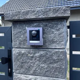 Zamontowany wideodomofon IP Hikvision na szarym, kamiennym słupku ogrodzenia, widoczny fragment furtki i domu z szarą dachówką w tle.