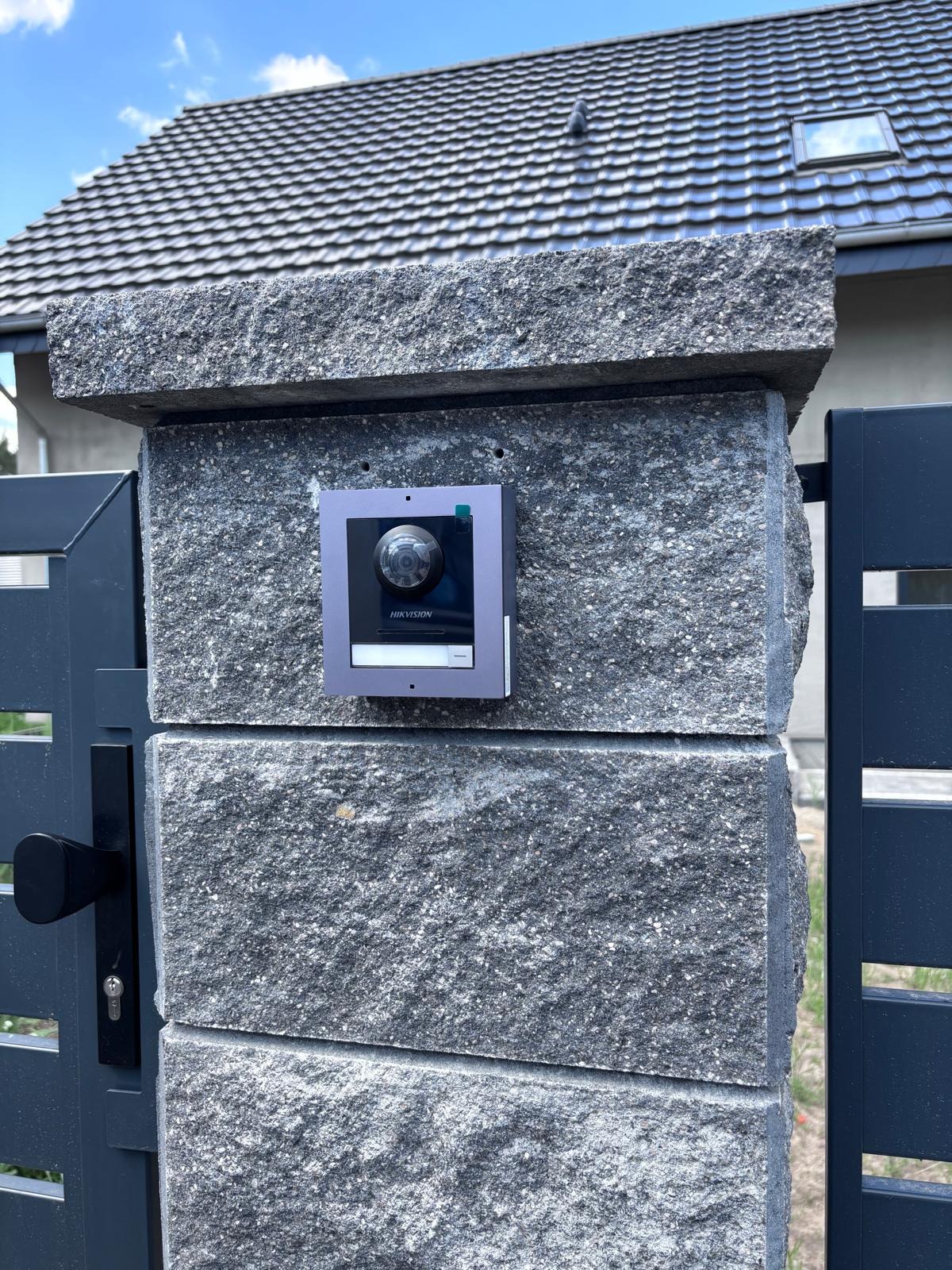 Zamontowany wideodomofon IP Hikvision na szarym, kamiennym słupku ogrodzenia, widoczny fragment furtki i domu z szarą dachówką w tle.