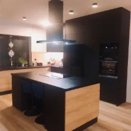 Nowoczesna kuchnia z wyspą, fronty szafek w kolorze jasnego drewna i czerni, czarny okap, oświetlenie punktowe, podłoga drewniana, minimalistyczny styl.