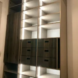 Wysoka szafa wnękowa z szarymi frontami, oświetleniem LED wewnątrz, widoczne półki i szuflady z ciemnymi uchwytami, drewniana podłoga.