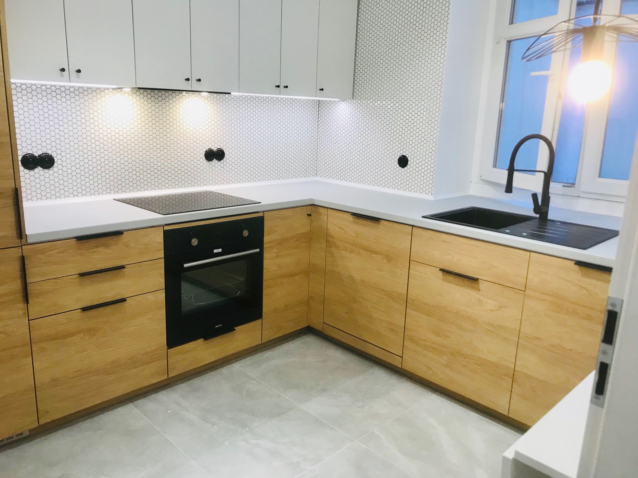 Nowoczesna kuchnia w jasnych barwach z drewnianymi frontami szafek, białymi górnymi szafkami, czarnym zlewem i baterią, oraz mozaiką heksagonalną na ścianie nad blatem.