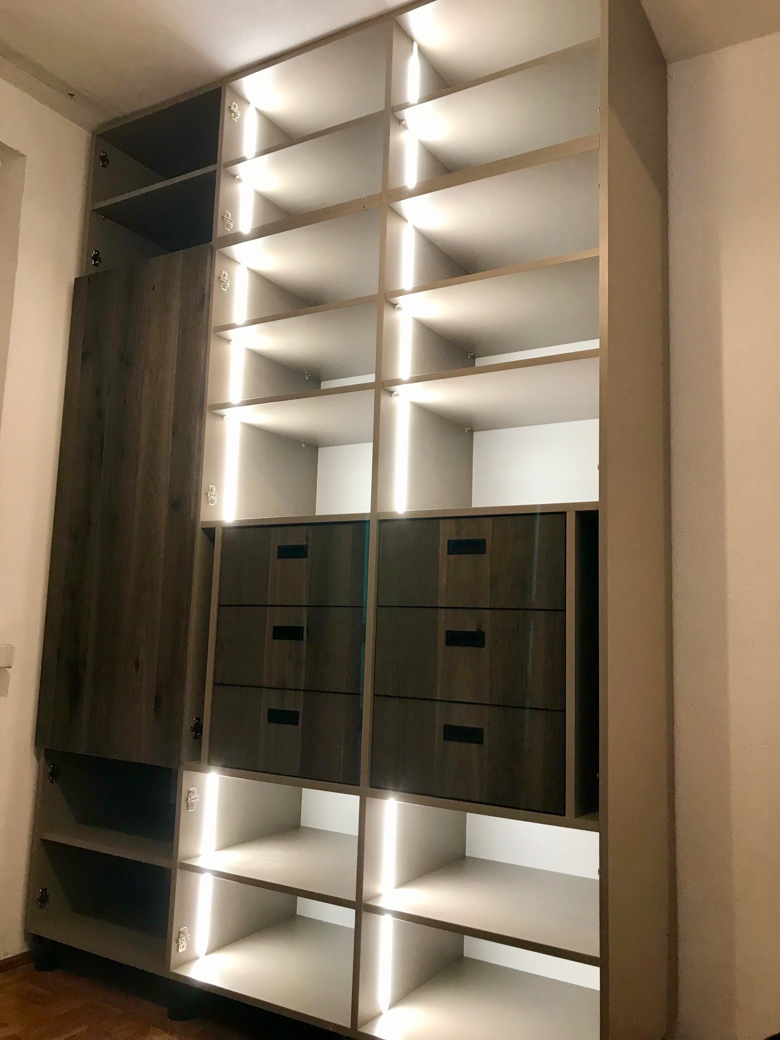 Wysoka szafa wnękowa z szarymi frontami, oświetleniem LED wewnątrz, widoczne półki i szuflady z ciemnymi uchwytami, drewniana podłoga.