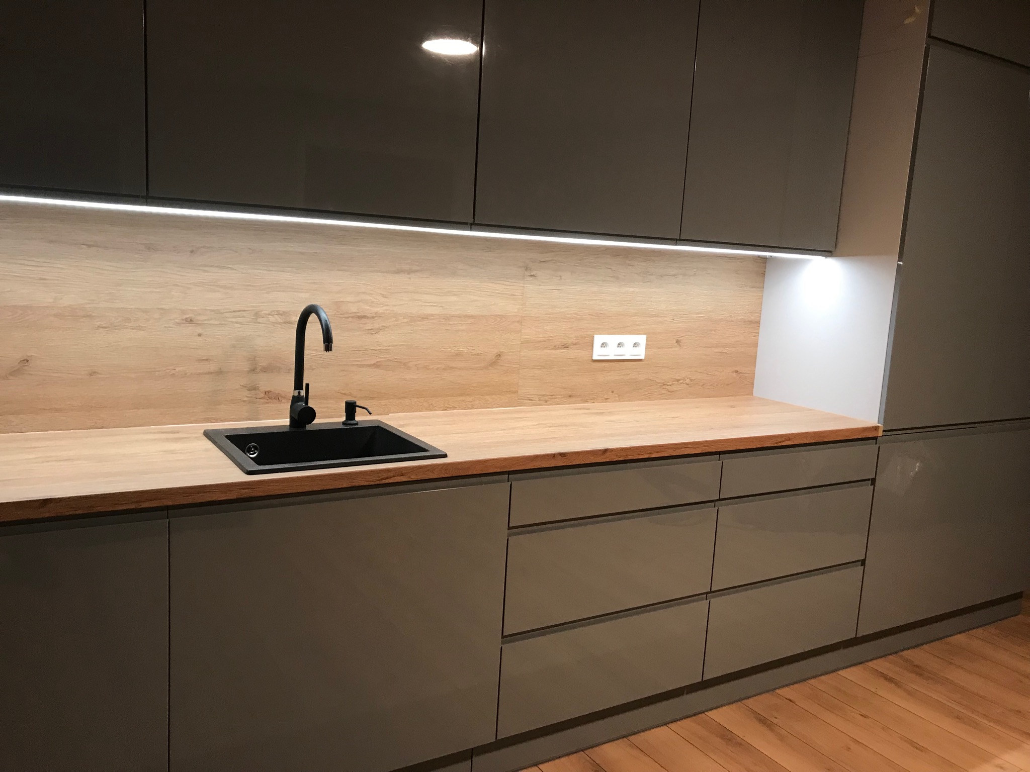 Nowoczesna kuchnia z szarymi, lakierowanymi frontami, drewnianym blatem, czarnym zlewem i baterią oraz oświetleniem LED podszafkowym.