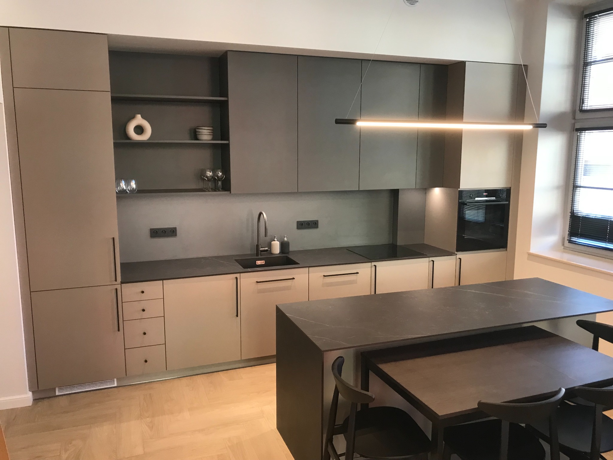 Nowoczesna kuchnia z szarymi frontami, wyspą z blatem imitującym kamień, oświetleniem liniowym LED i drewnianą podłogą w jodełkę.