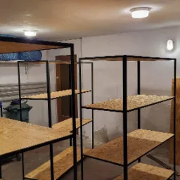 Metalowe regały z drewnianymi półkami OSB, czarne ramy, surowe wnętrze magazynu lub piwnicy, widoczne ślady wilgoci na ścianach, oświetlenie sufitowe