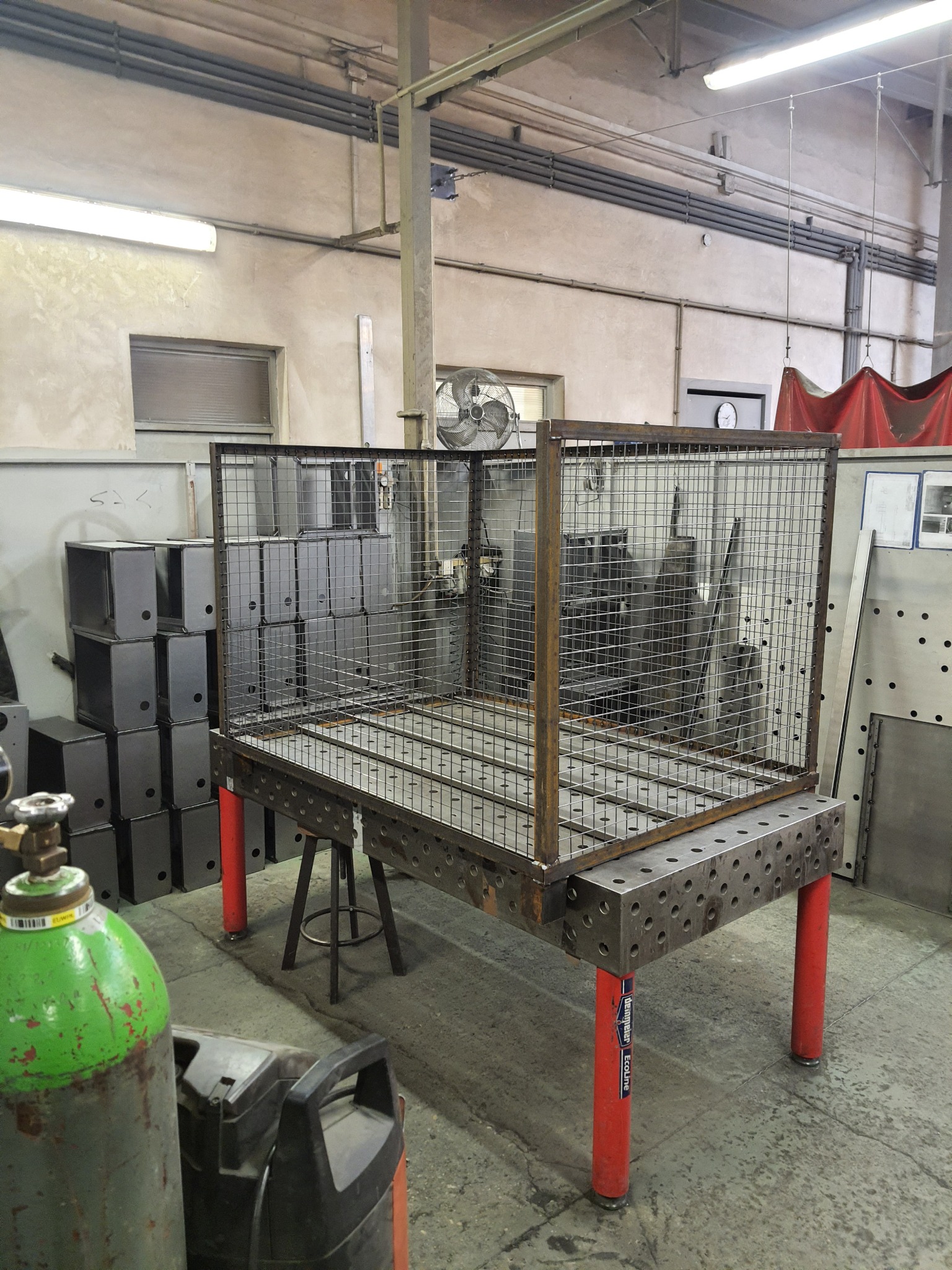 Klatka z metalowej siatki spawana na stole spawalniczym z czerwonymi nogami, w tle widoczne metalowe szafki i butla z gazem.