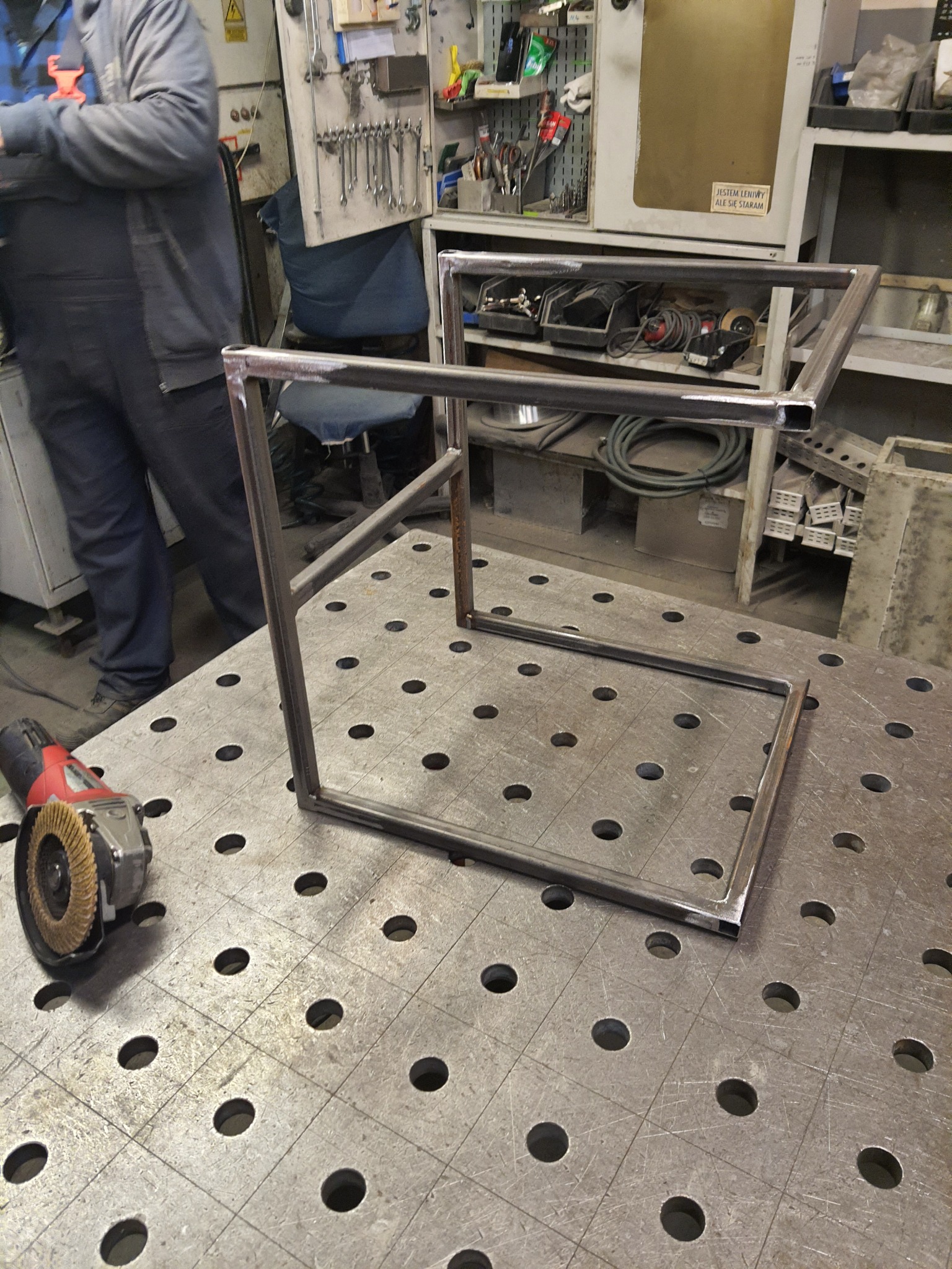 Metalowa konstrukcja z profili kwadratowych, częściowo pospawana, leżąca na stole spawalniczym z perforacją, obok szlifierka kątowa z tarczą lamelkową, w tle warsztat.