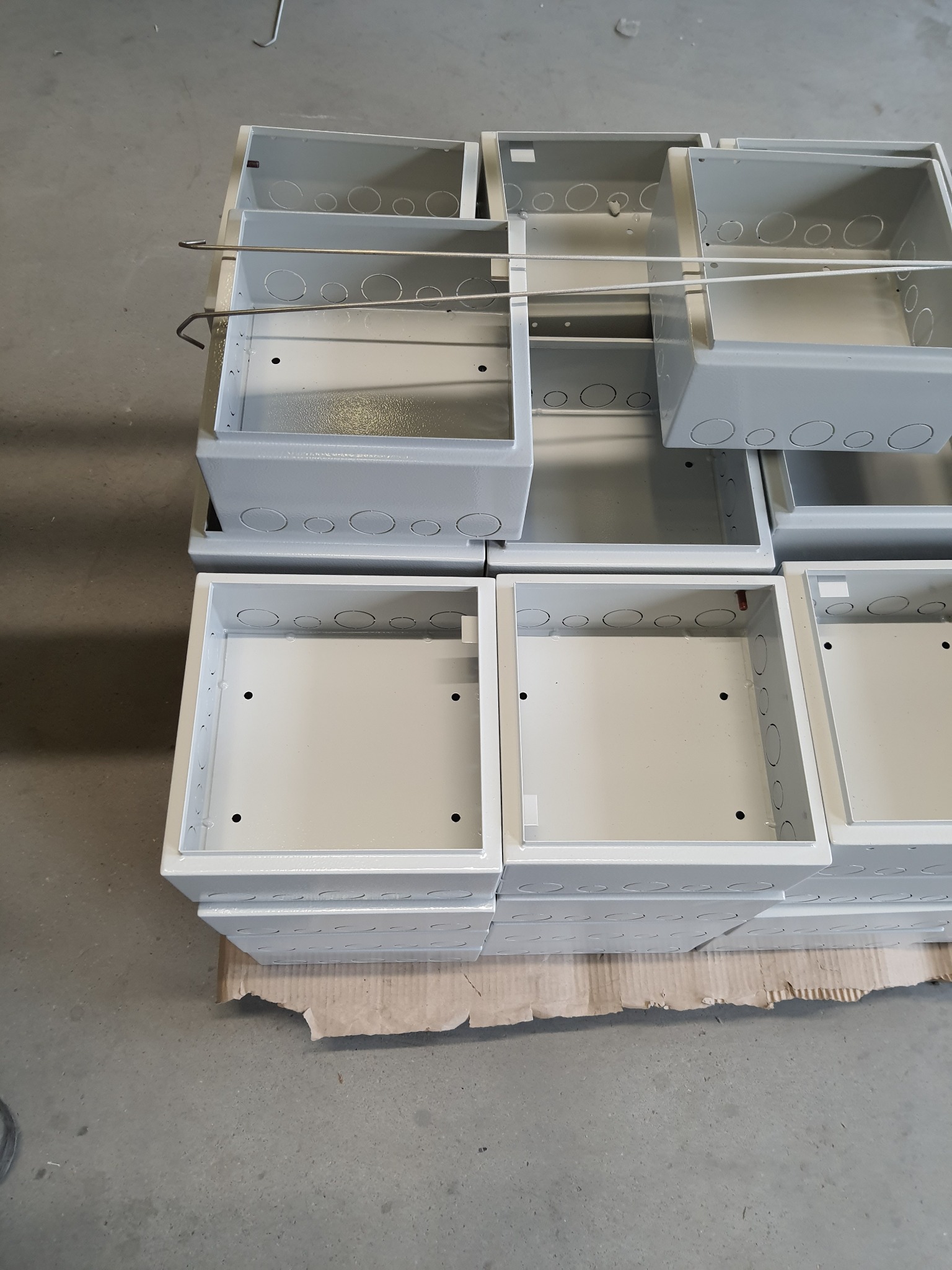 Stos szarych, metalowych obudów elektrycznych z otworami na kable, ułożonych na kartonie na betonowej podłodze.