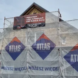 Budynek murowany w trakcie prac elewacyjnych, osłonięty siatką z logo Atlas, widoczne rusztowania i baner firmy budowlanej.