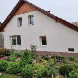 Świeżo otynkowany dom murowany z drewnianą podbitką, widok z ogrodu z bujną roślinnością i elementami małej architektury.