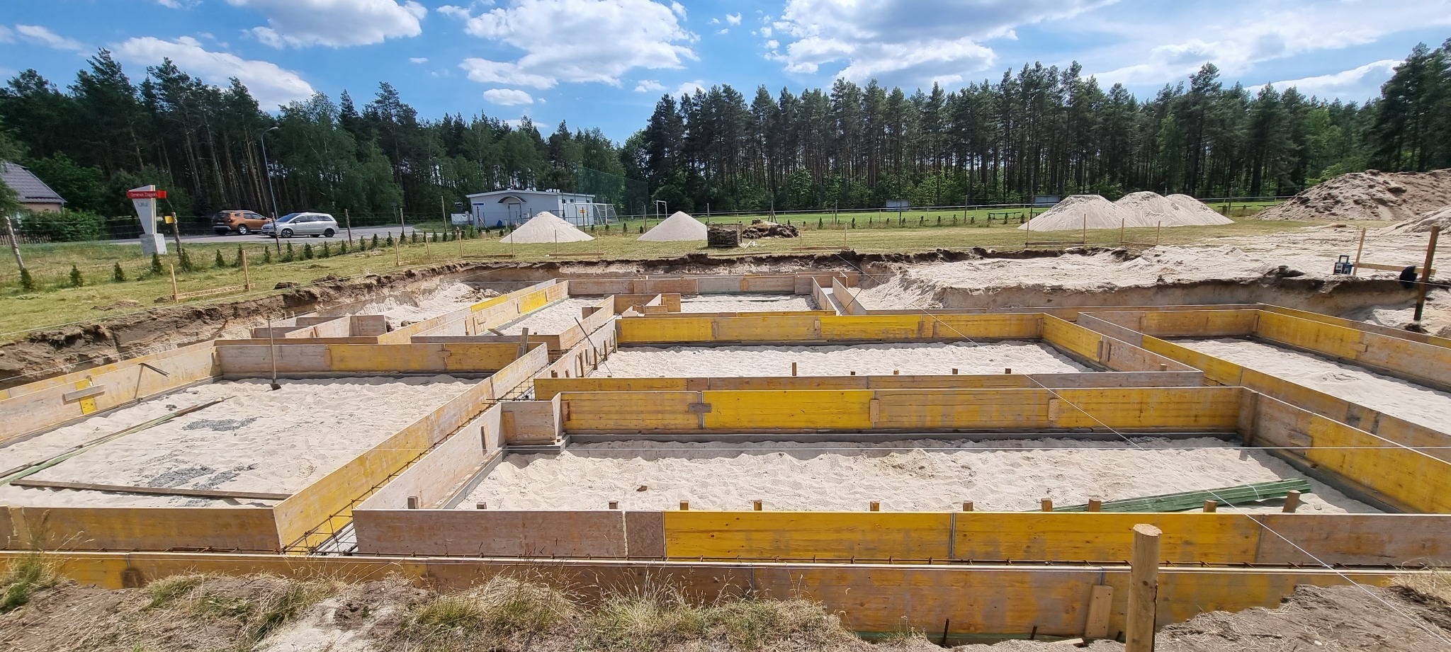 Przygotowanie szalunków pod fundamenty domu jednorodzinnego, wypełnione piaskiem, widok z góry na wykop z żółtymi deskami, w tle hałdy piachu i zalesiony teren.