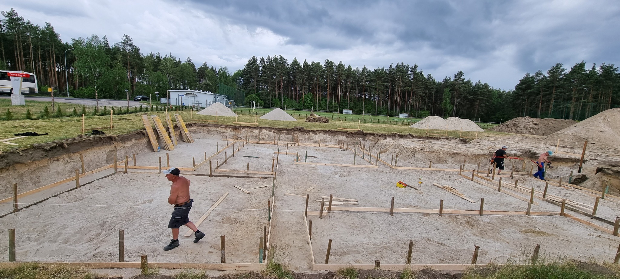 Przygotowanie terenu pod fundamenty domu: wykopany dół z drewnianym szalunkiem, widoczne pryzmy piasku i dwóch pracowników z narzędziami, tło stanowią drzewa i pochmurne niebo.