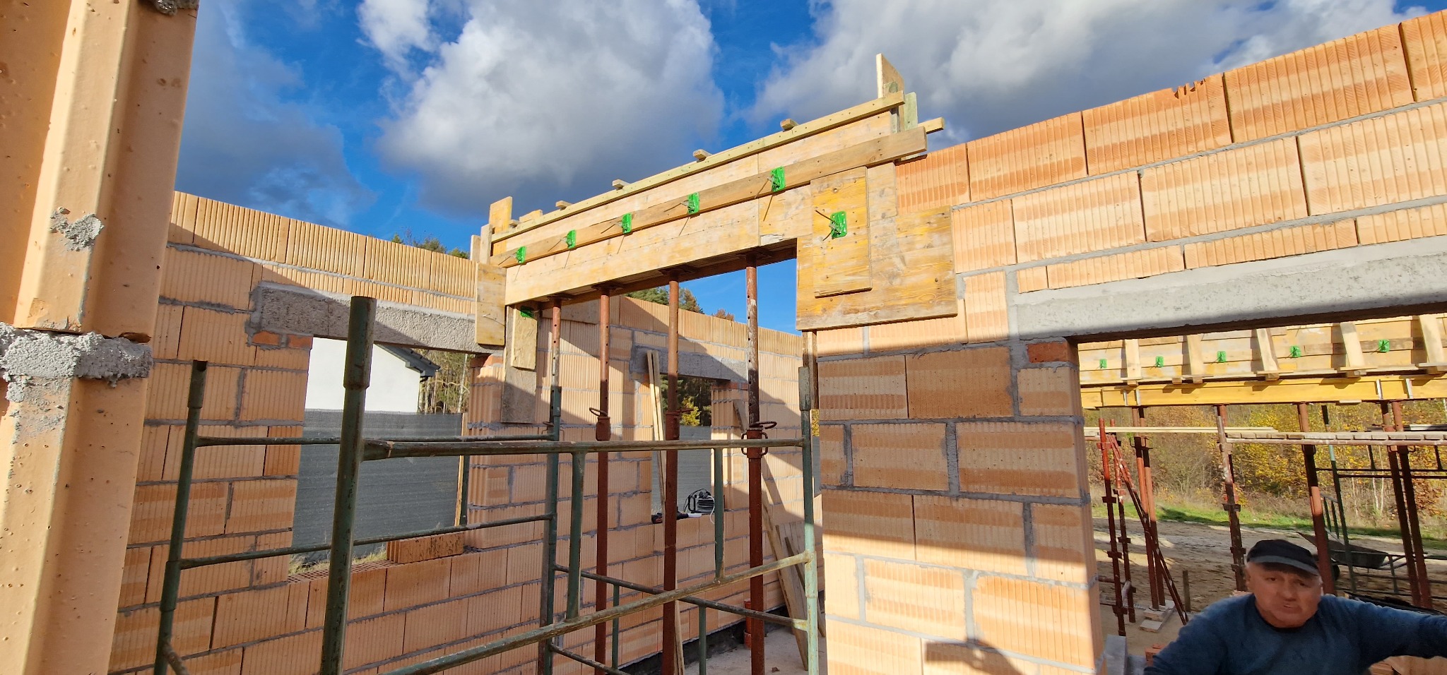 Wznoszenie ścian budynku z bloczków ceramicznych, widoczne drewniane szalunki nad otworami okiennymi i drzwiowymi, stalowe podpory i rusztowania, niebo z chmurami w tle.