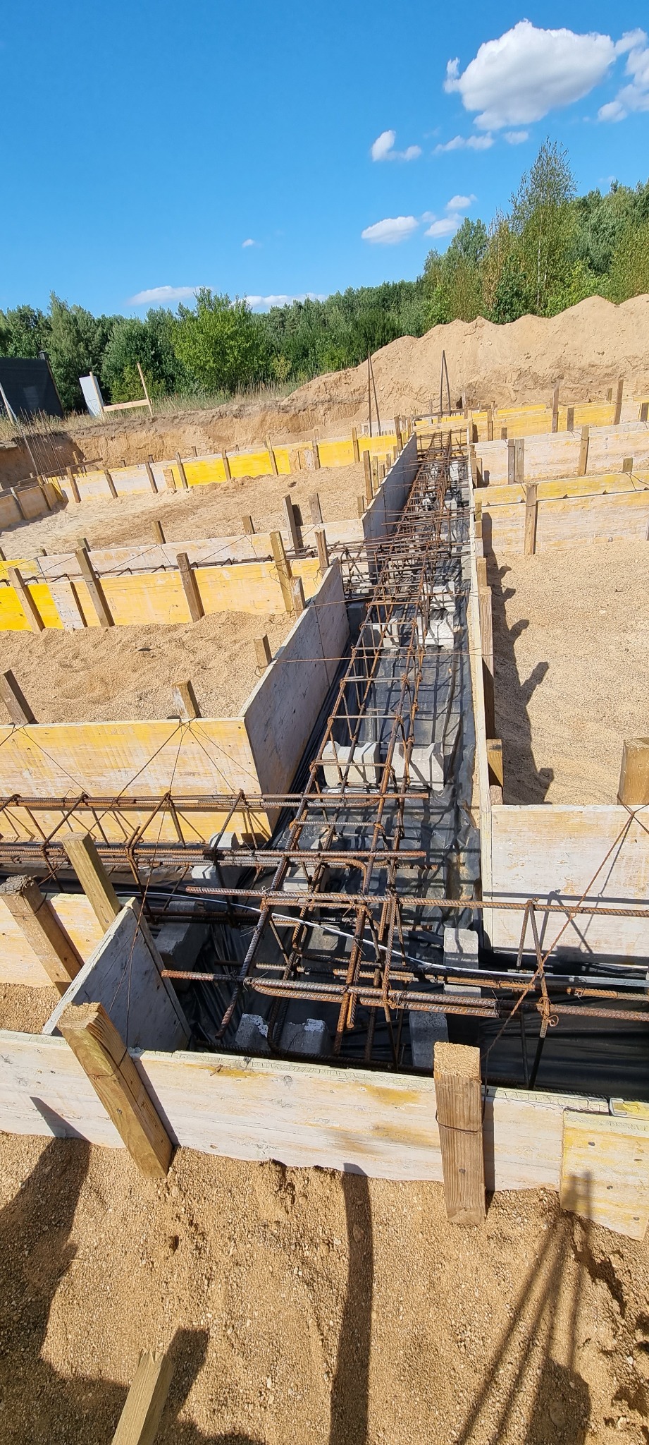 Zbrojenie w szalunku fundamentowym, widok z góry na przygotowany wykop z drewnianym szalunkiem i siatką zbrojeniową przed zalaniem betonem, w tle usypana ziemia i drzewa pod błękitnym niebem.
