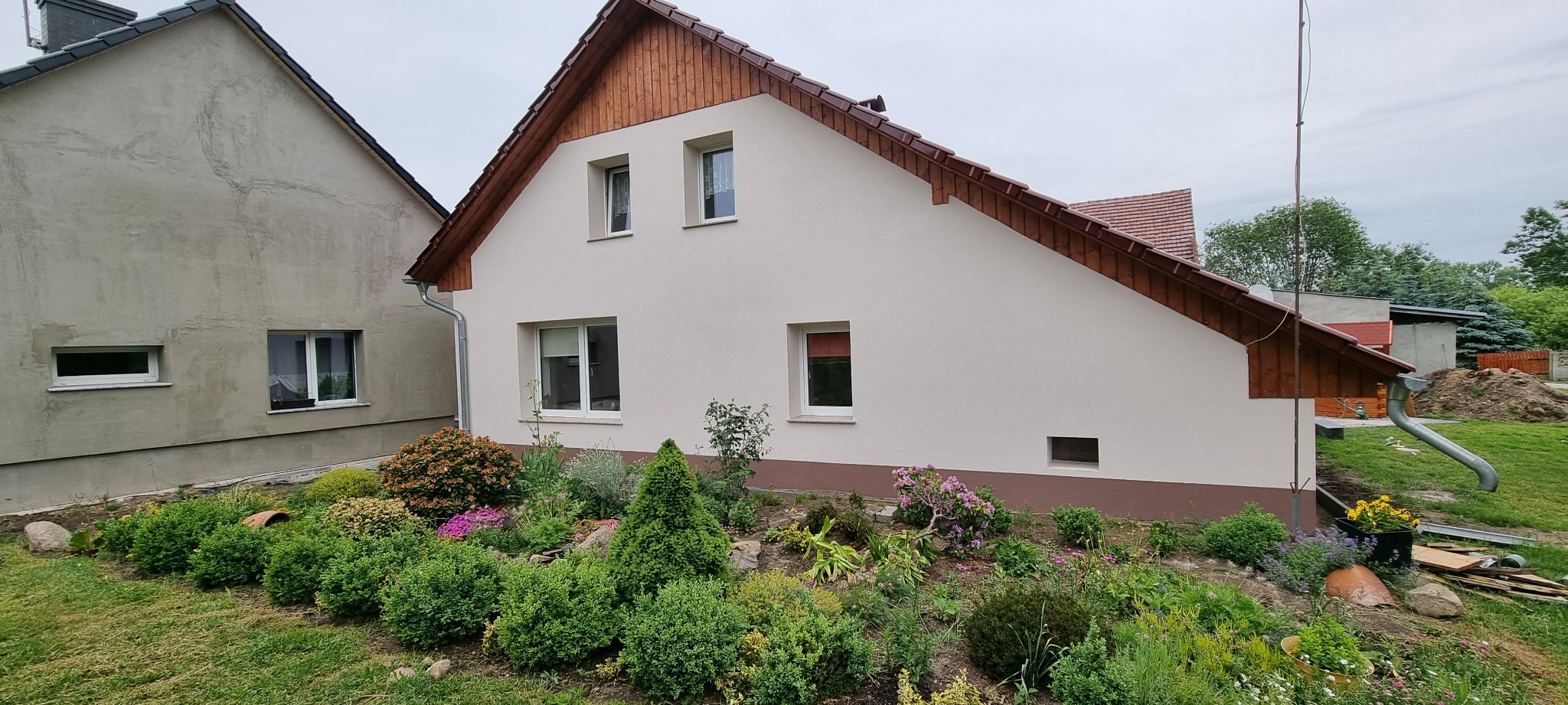 Świeżo otynkowany dom murowany z drewnianą podbitką, widok z ogrodu z bujną roślinnością i elementami małej architektury.