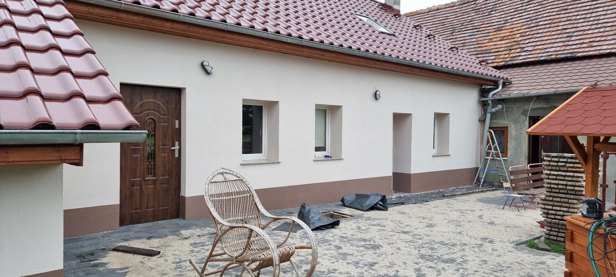 Świeżo otynkowany dom murowany z brązowym pasem u dołu, widoczne okna, drzwi wejściowe i wiklinowy fotel na pierwszym planie, teren wokół domu wyłożony kostką.