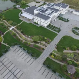 Widok z lotu ptaka na rozległy teren z budynkiem, parkingiem i starannie zagospodarowanym ogrodem z elementami małej architektury, w tym ścieżki z kostki, mostek i plac zabaw.