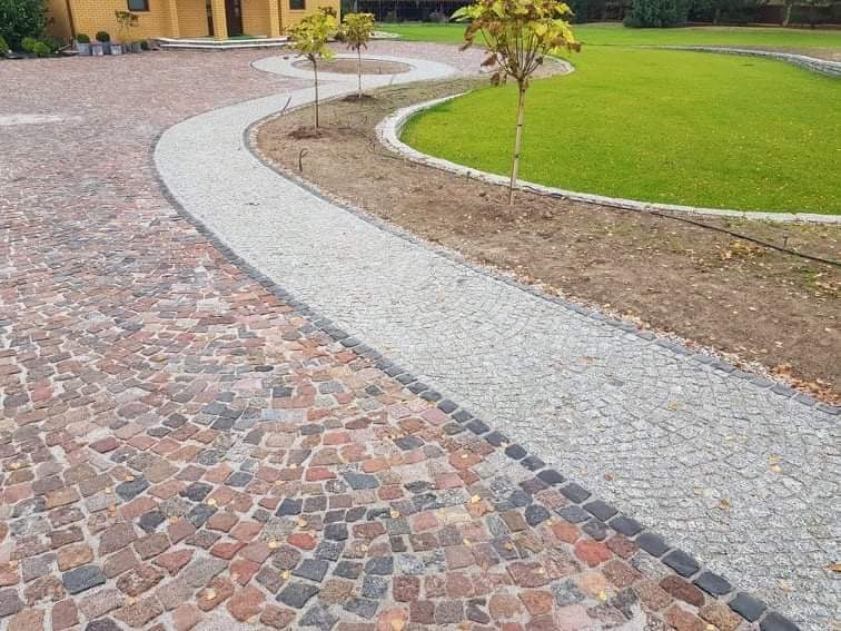 Szeroki podjazd z kostki granitowej w dwóch kolorach: czerwono-brązowym i szarym, oddzielone pasem czarnej kostki. Szara ścieżka prowadzi do trawnika z młodymi drzewkami.