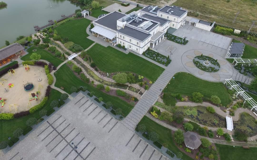 Widok z lotu ptaka na rozległy teren z budynkiem, parkingiem i starannie zagospodarowanym ogrodem z elementami małej architektury, w tym ścieżki z kostki, mostek i plac zabaw.