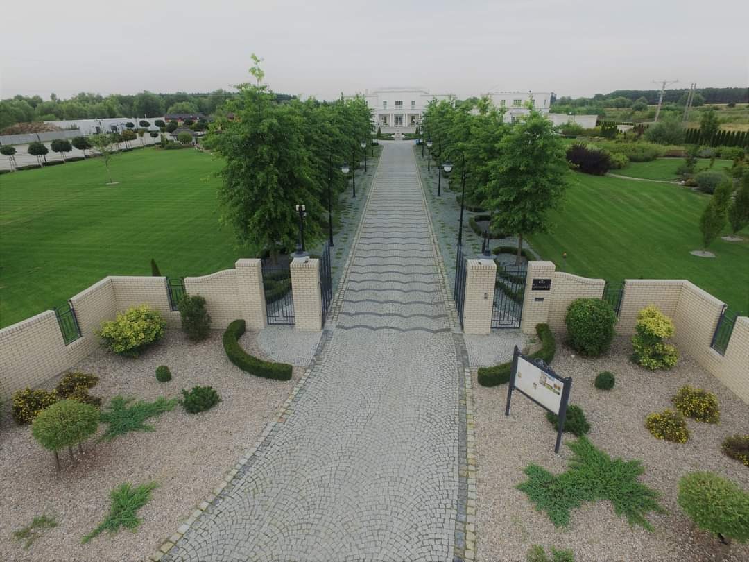 Szeroki podjazd z jasnej kostki prowadzący do białego budynku, otoczony równo przystrzyżonym trawnikiem, niskimi murkami z jasnej cegły i ozdobnymi krzewami.