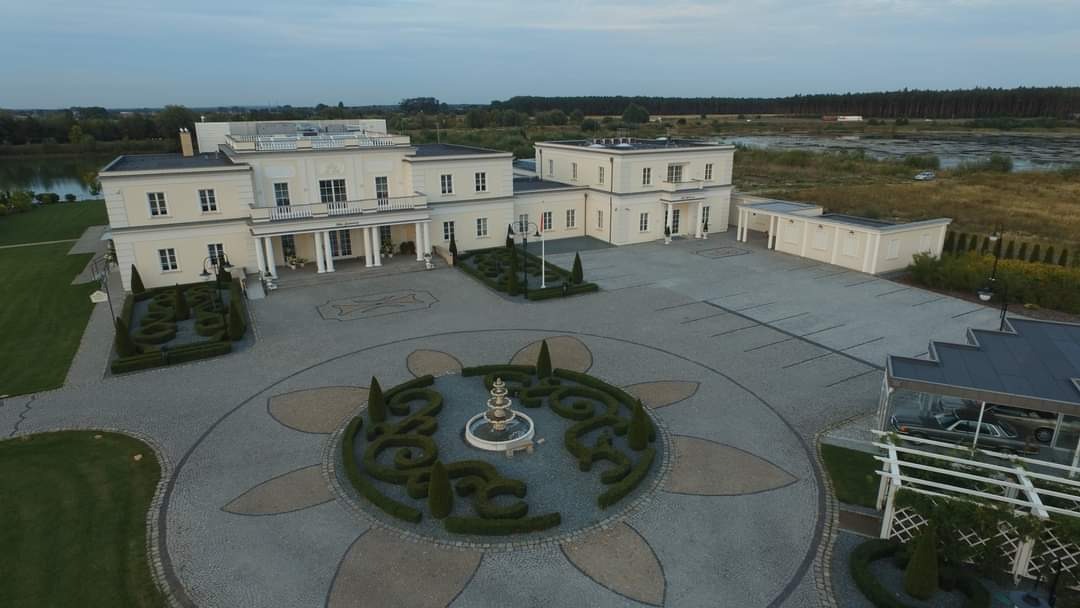 Rozległy dziedziniec rezydencji z fontanną otoczoną ozdobnymi krzewami i kostką granitową ułożoną we wzór kwiatowy, widok z lotu ptaka.