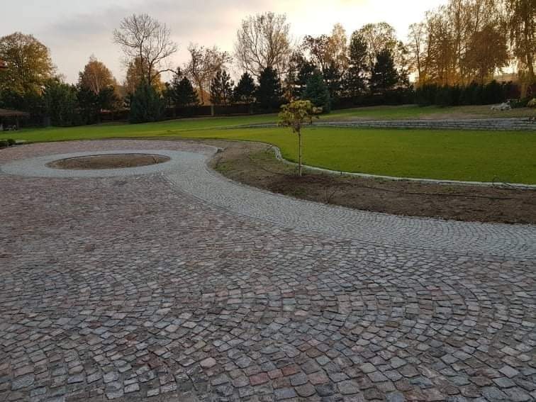 Szeroka, wybrukowana droga z kostki w kształcie wachlarza prowadząca przez zadbany trawnik z młodym drzewkiem w centrum okręgu, otoczona zielenią i drzewami w tle.