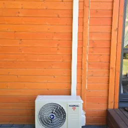 Zewnętrzna jednostka klimatyzacji Kaisai zamontowana na czarnych wspornikach przy drewnianej ścianie domu, z widoczną białą rurą odprowadzającą i kostką brukową.