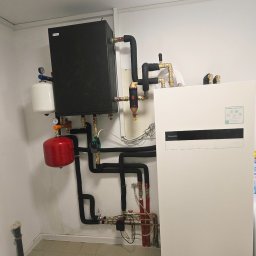 Hydro Gorzów - Zestaw instalacji grzewczej z czarnymi rurami, czerwonym zbiornikiem i białym panelem Panasonic Aquarea Smart Cloud na tle białej ściany i beżowej podłogi.