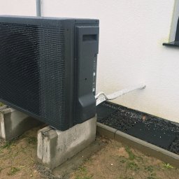 Hydro Gorzów - Czarna pompa ciepła Aquarea Panasonic na betonowych podstawach, zamontowana przy białej ścianie domu z oknem. Widoczne rury odprowadzające.